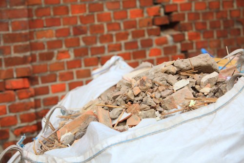 A large white flexible construction waste bag filled with a mixture of broken bricks, concrete fragments, and small pieces of rubble, situated outdoors against a red brick wall. The rubble appears uneven and varied in size, with some larger pieces and smaller debris scattered throughout. The bag is partially open at the top, revealing the debris inside, and secured with blue ties on the sides. The surrounding environment suggests an external area, possibly a driveway or back alley in a residential or commercial setting, relevant to rubbish removal services provided by Office Clearance Catford in the London borough, near Catford. The lighting indicates natural daylight, highlighting the textures of the debris and brick surface, aligning with typical waste collection or clearance activities in the area.
