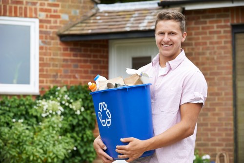 A young man with short, light brown hair and fair skin is standing outdoors in front of a brick house with a tiled roof and a large window. He is smiling and wearing a light pink, short-sleeved button-up shirt. The man is holding a blue recycling bin with a white recycling symbol on the side, filled with various waste items such as paper, cardboard, and plastic bottles, which are visible at the top of the bin. Behind him, there is a green shrub with white flowering clusters, and the background includes a portion of a white doorframe and a pathway leading to the house. The scene is well-lit with natural daylight, typical of a residential environment, suggesting a rubbish collection or waste disposal task possibly related to local clearing services in the Catford area. The image reflects a professional approach to rubbish removal, highlighting the importance of proper waste separation and collection practices in the community served by Office Clearance Catford.