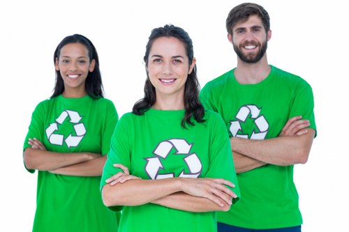 Three individuals wearing bright green T-shirts featuring a white recycling symbol stand outdoors against a neutral background. The woman in the center has shoulder-length dark hair and is smiling directly at the camera, with her arms crossed in front of her. To her left, a woman with long dark hair is also smiling and has her arms crossed, while to her right, a man with short dark hair and a beard is smiling broadly and has his arms crossed as well. The group appears to be part of a rubbish removal or recycling initiative, supporting environmental sustainability efforts. Their clothing and confident posture suggest they are ready to assist with waste and rubbish clearance services, possibly in the Catford area. The setting is outdoors, illuminated by natural light, and their presence visually emphasizes the importance of responsible waste management and recycling, aligning with the services offered by Office Clearance Catford.