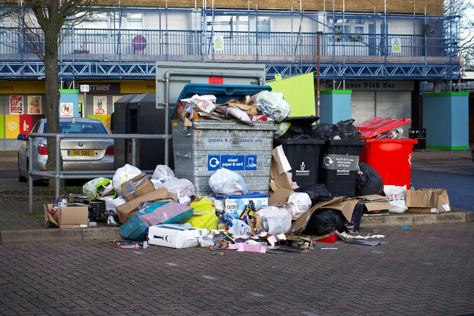 A large overflowing collection of rubbish situated on a paved sidewalk in front of a commercial area with multiple waste bins, some of which are black and others red, containing a mix of discarded car